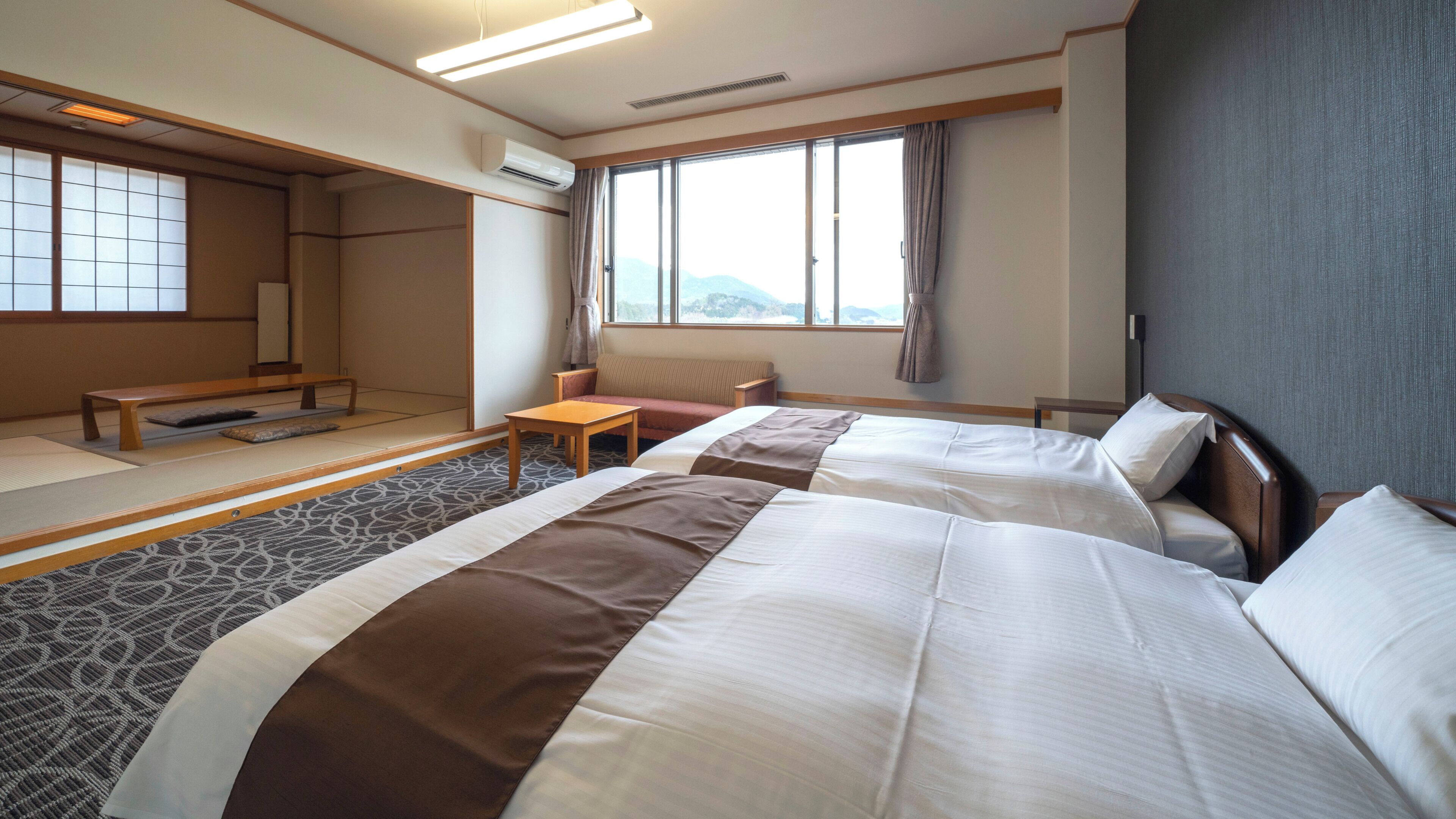 A hotel room with two beds, a small table, a sofa, a window with a view, and traditional Japanese tatami flooring.