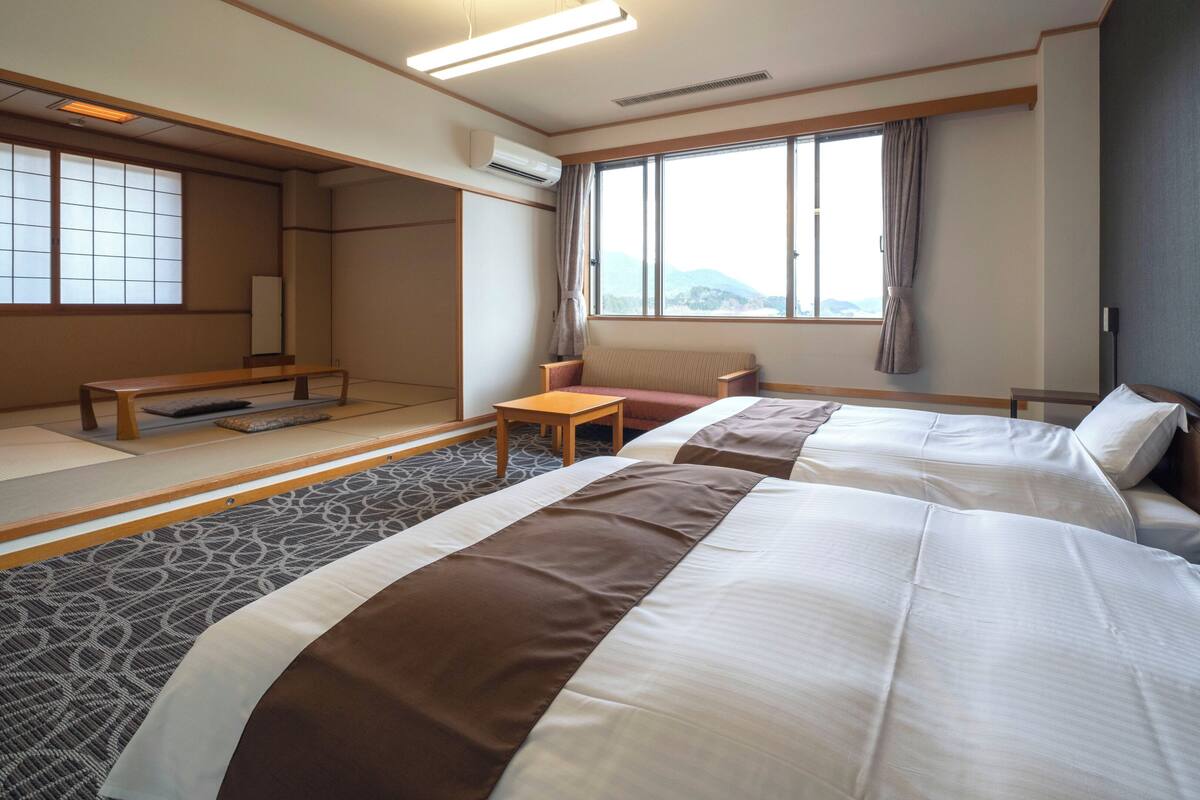 Habitación de hotel con dos camas, una mesita, un sofá, una ventana con vistas y suelo tradicional de tatami japonés.