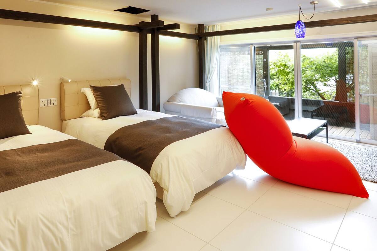 Habitación de hotel con dos camas, una silla roja y vista a zonas verdes.