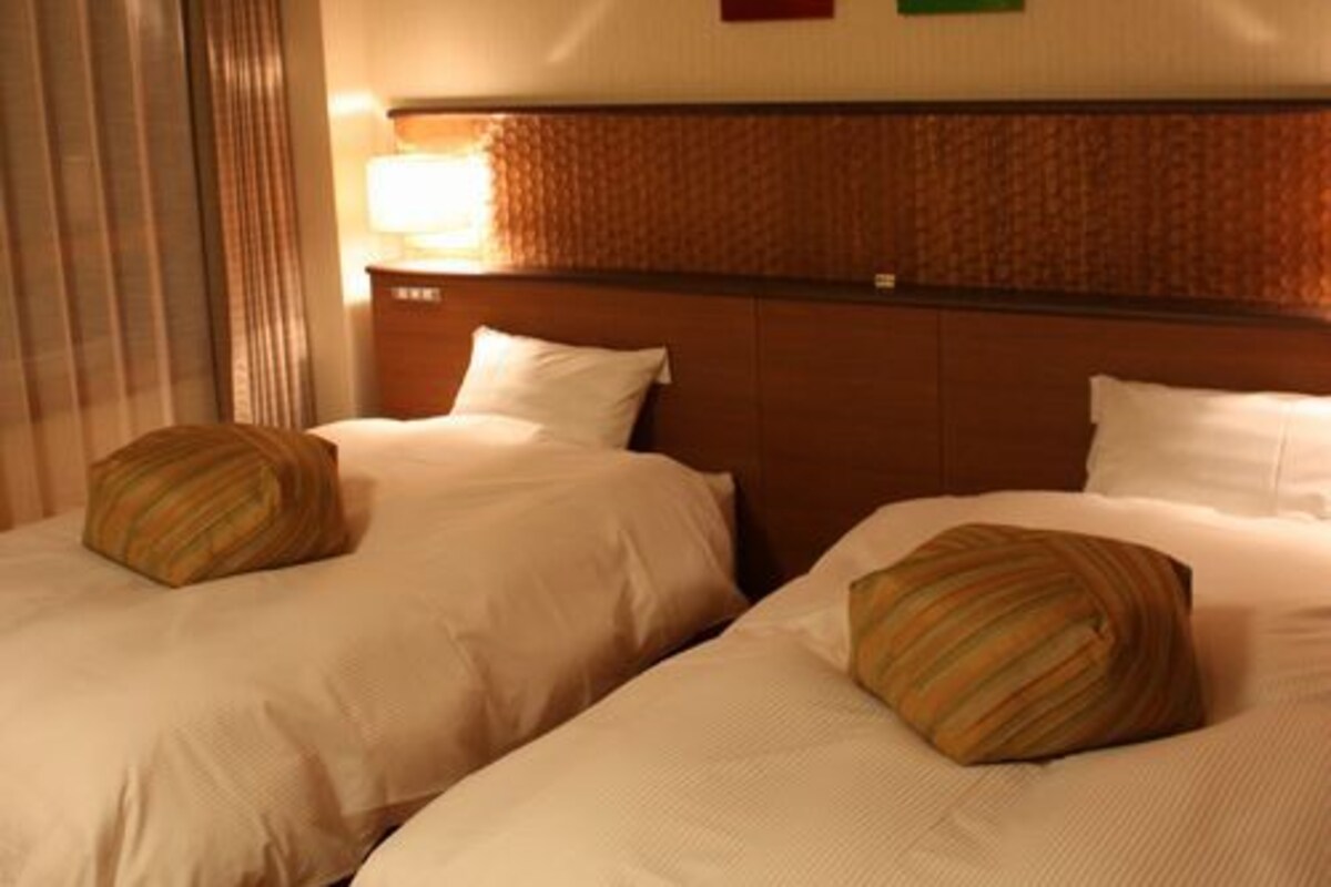 Ein Hotelzimmer mit zwei Betten, einem Kopfteil, einem Fenster mit Vorhängen und einer Wand mit zwei farbigen Paneelen.