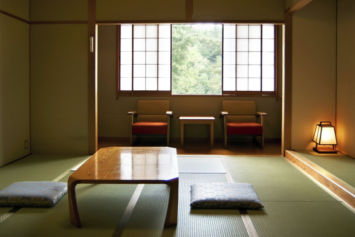 Une pièce de style japonais traditionnel, avec un sol recouvert de tatamis, une table basse, deux chaises et une fenêtre donnant sur un paysage verdoyant.