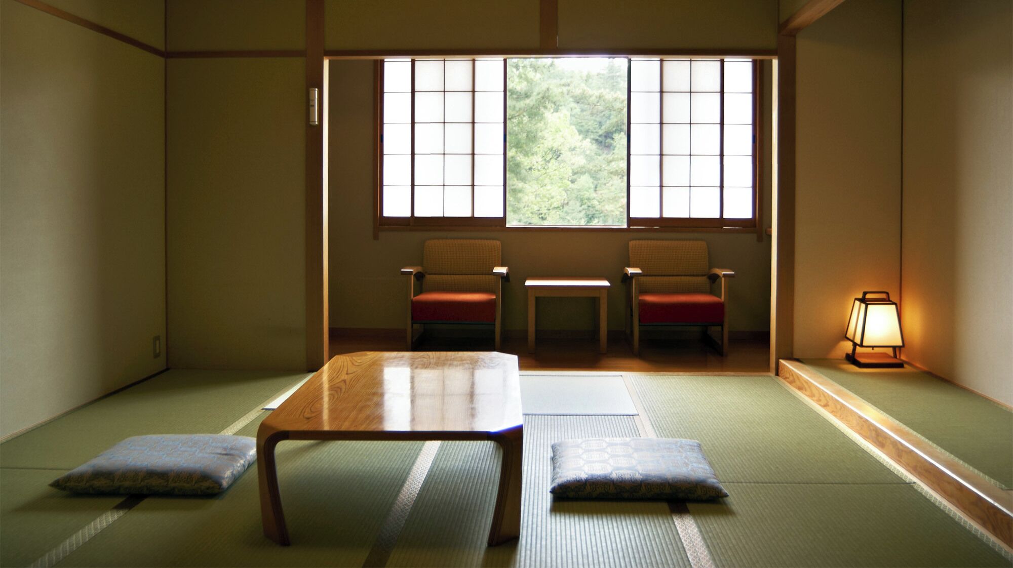 A traditional Japanese room with tatami flooring, a low table, two chairs, and a window with a view of greenery.