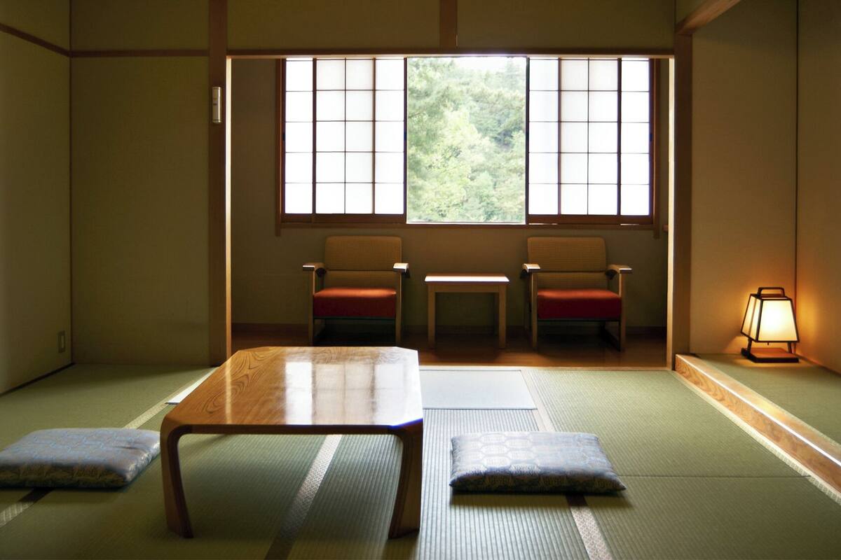 Una habitación japonesa tradicional con suelo de tatami, una mesa baja, dos sillas y una ventana con vistas a la vegetación.