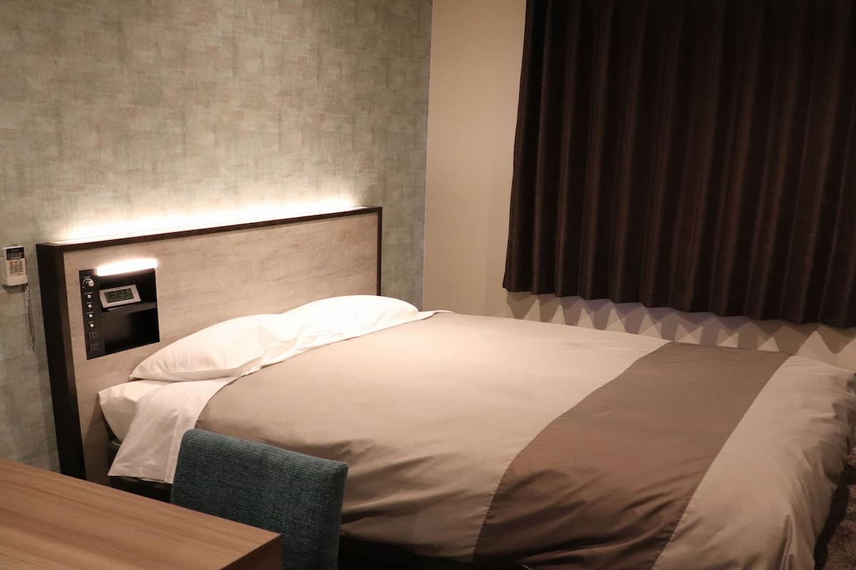 A hotel room with a bed, a bedside table, a phone, and a window with curtains.
