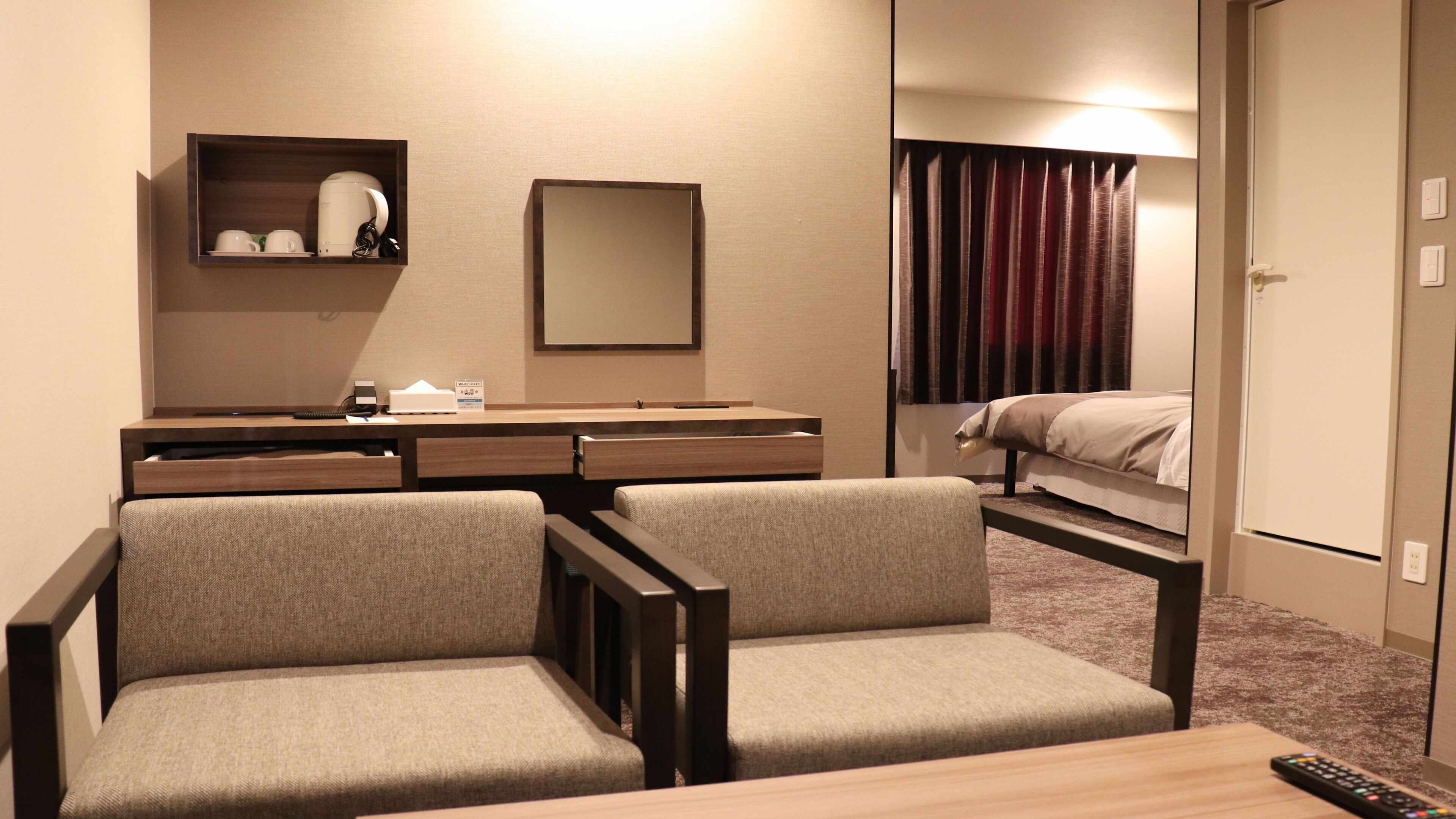 A hotel room with a desk, two chairs, a bed, a mirror, and a window with curtains.