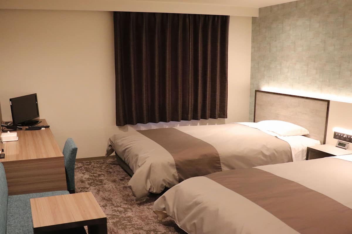 A hotel room with two beds, a desk with a computer, a chair, and a large window with curtains.