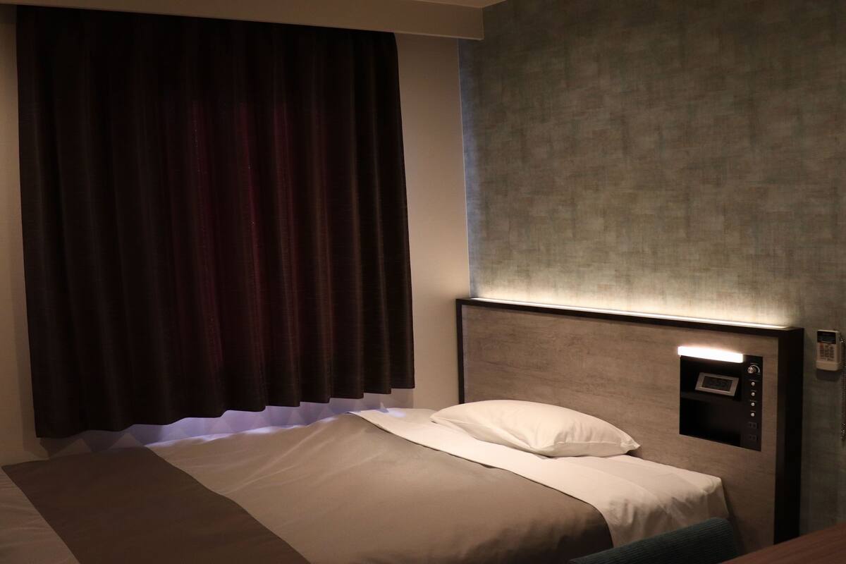 A hotel room with a bed, a nightstand, a wall-mounted phone, and a window with curtains.