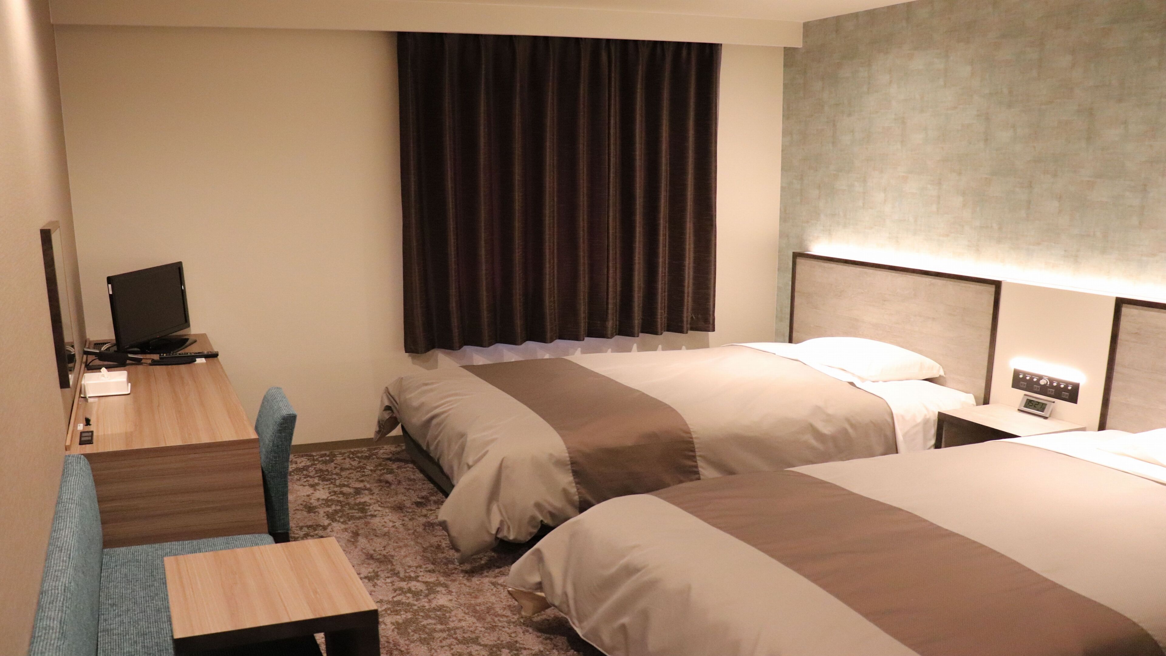A hotel room with two beds, a desk with a computer, a chair, and a large window with curtains.