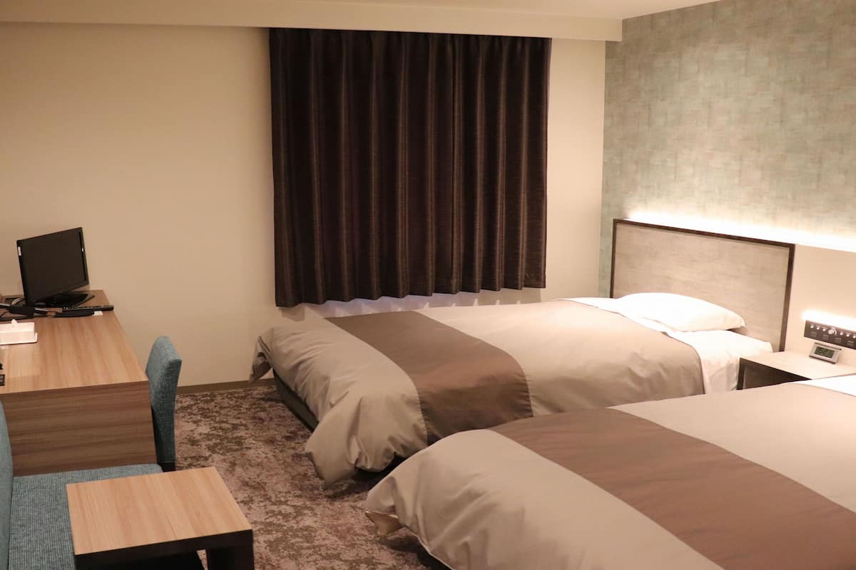 A hotel room with two beds, a desk with a computer, a chair, and a large window with curtains.