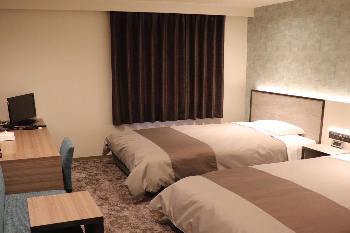 A hotel room with two beds, a desk with a computer, a chair, and a large window with curtains.
