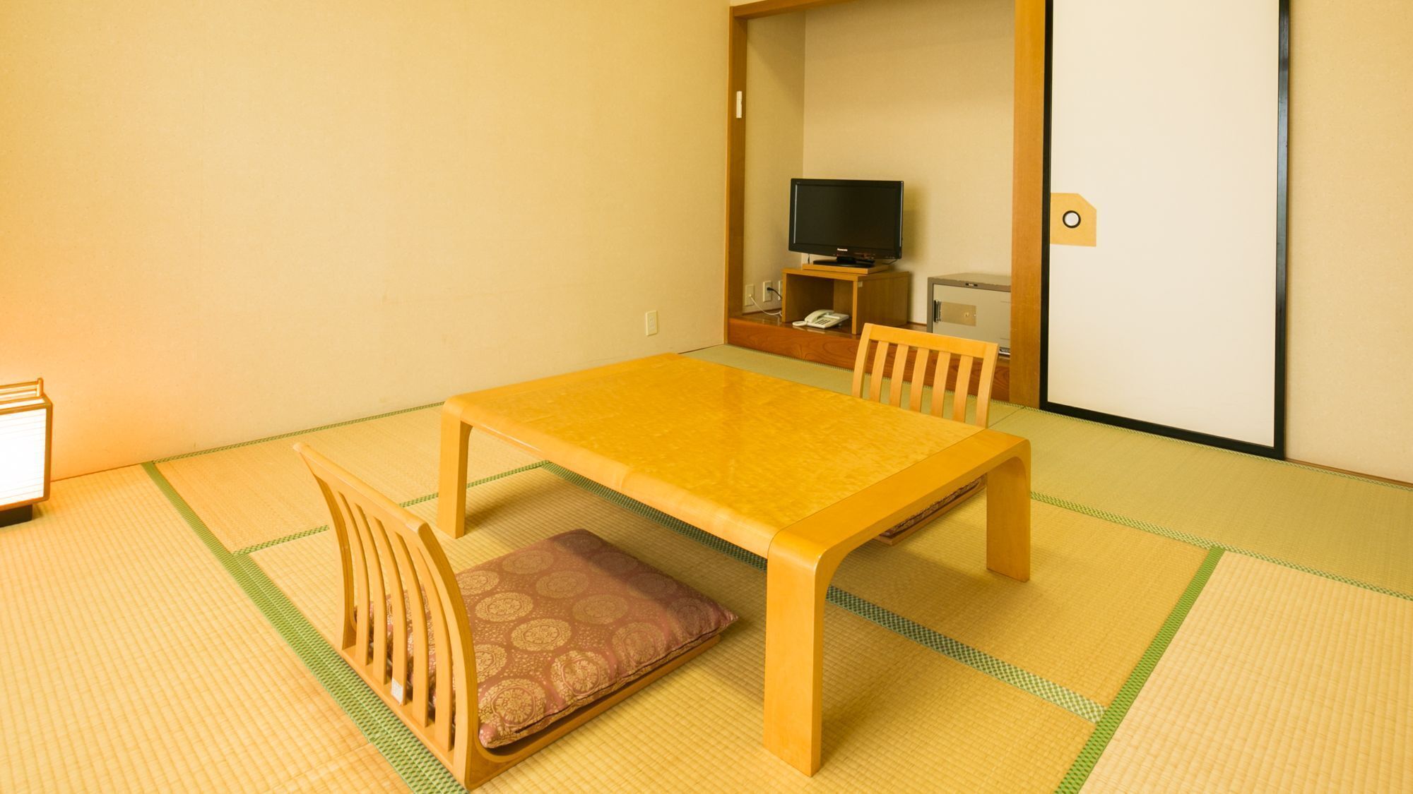 Ein Zimmer mit Tatami-Bodenbelag, einem Holztisch mit Kissen, einem Stuhl, einem Fernseher und einem Schrank.