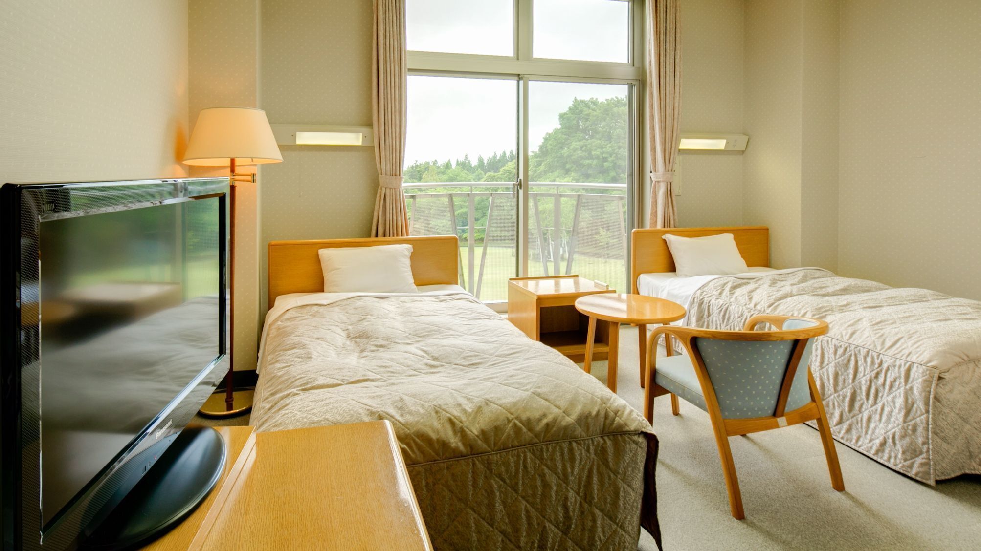 Ein Hotelzimmer mit zwei Betten, einem Fernseher, einem Schreibtisch, einem Stuhl und einem Fenster mit Blick auf Grünflächen.