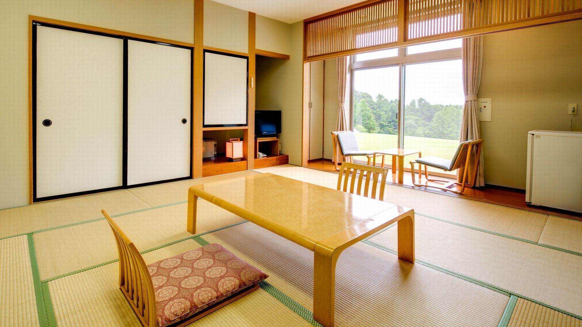 Ein traditioneller japanischer Raum mit Tatami-Boden, einem niedrigen Tisch, Stühlen, einem Fernseher und Schiebetüren.