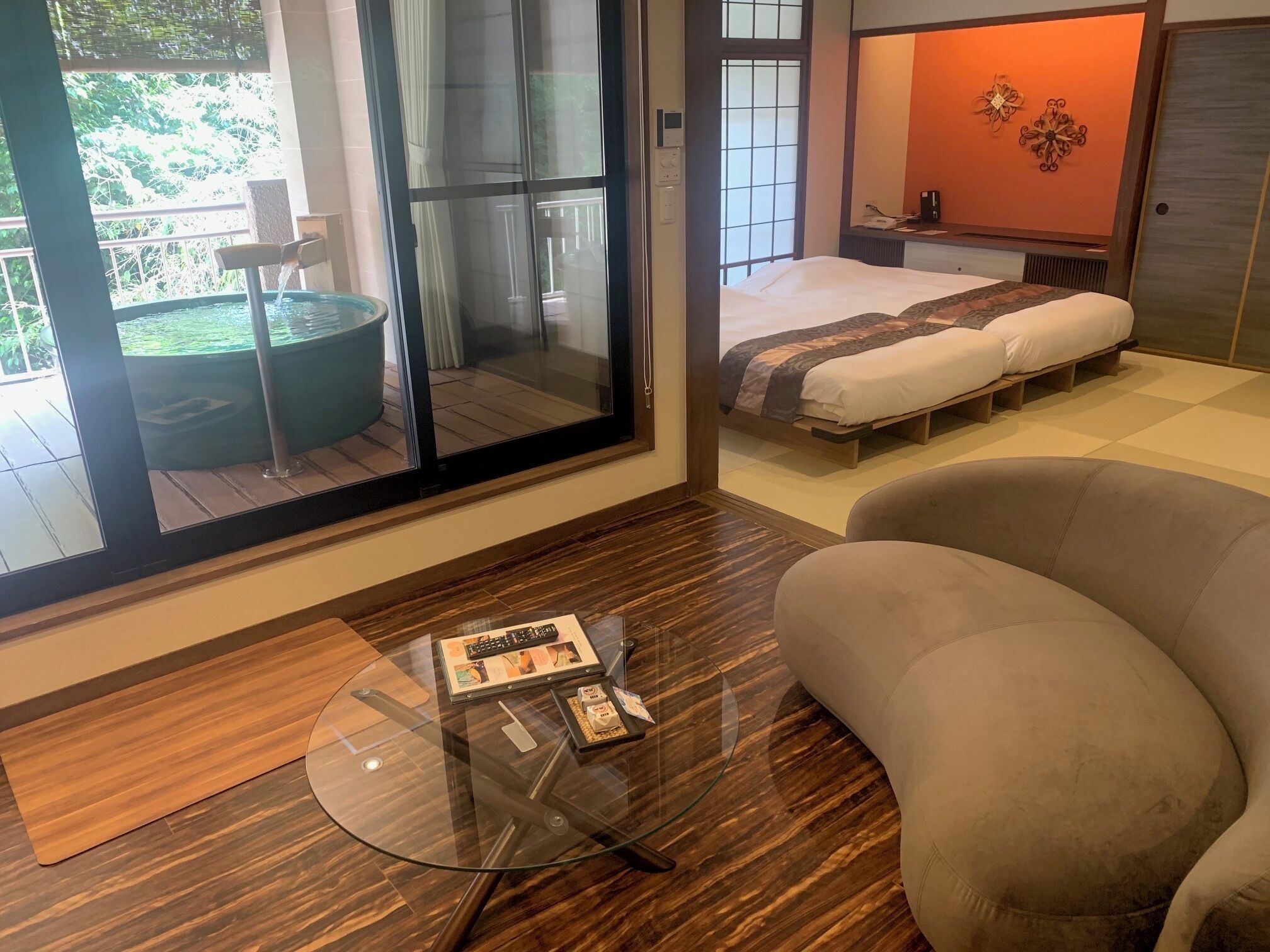 Ein modernes Schlafzimmer mit einem großen Schiebeglasfenster, das auf einen Balkon führt, einem Bett mit gemustertem Überwurf und einem Glastisch mit einer Zeitschrift.