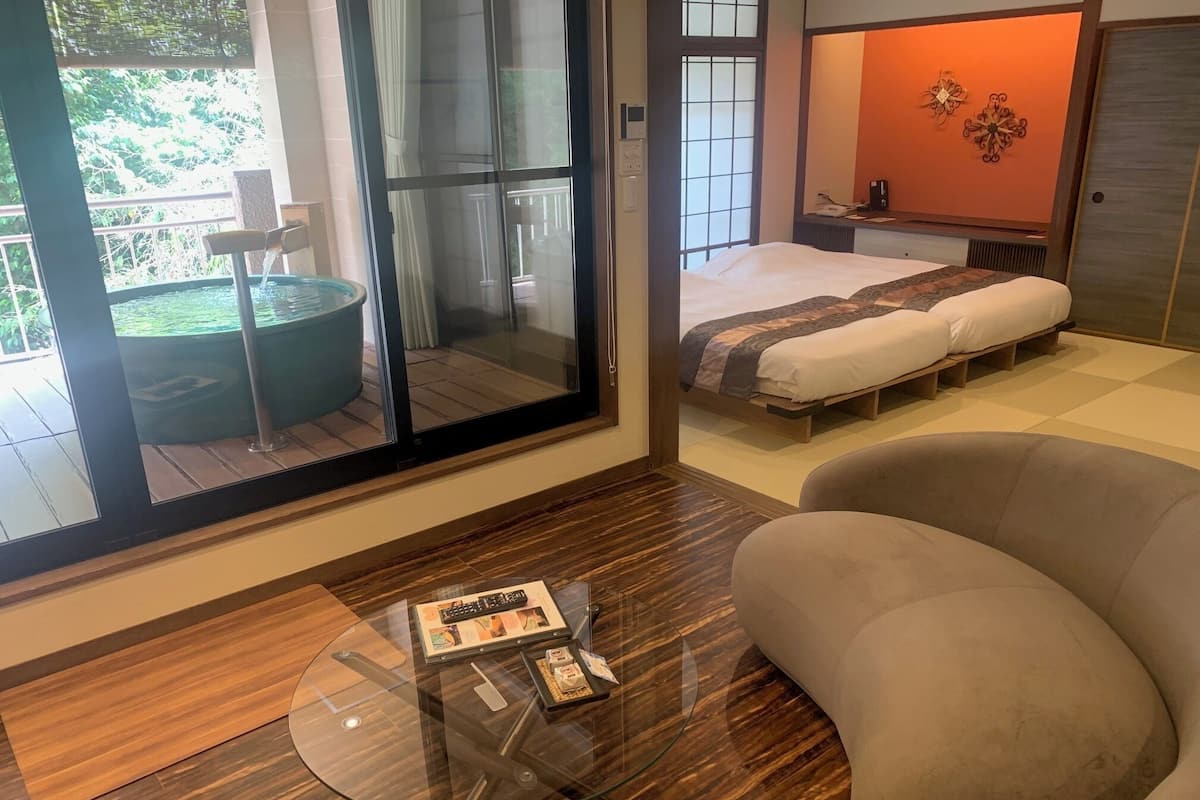 Un dormitorio moderno con un amplio ventanal corredero que da a un balcón, una cama con cubrecama estampado y una mesa de centro de vidrio con una revista.