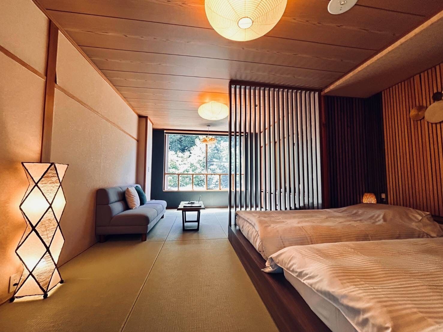 Ein modernes Schlafzimmer mit einer Couch, zwei Betten, einem kleinen Tisch und einem Fenster mit Blick auf Bäume.