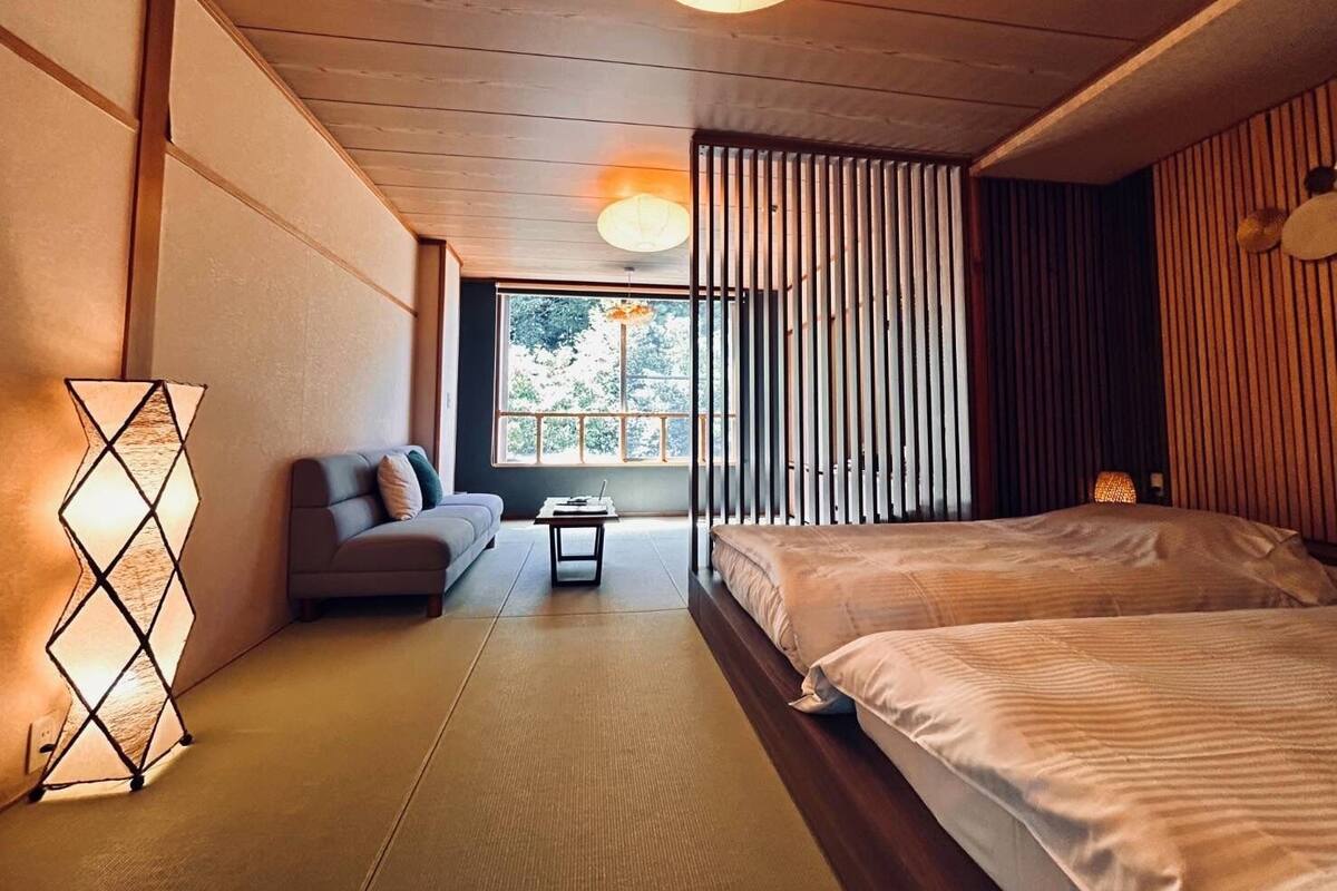 Ein modernes Schlafzimmer mit einer Couch, zwei Betten, einem kleinen Tisch und einem Fenster mit Blick auf Bäume.