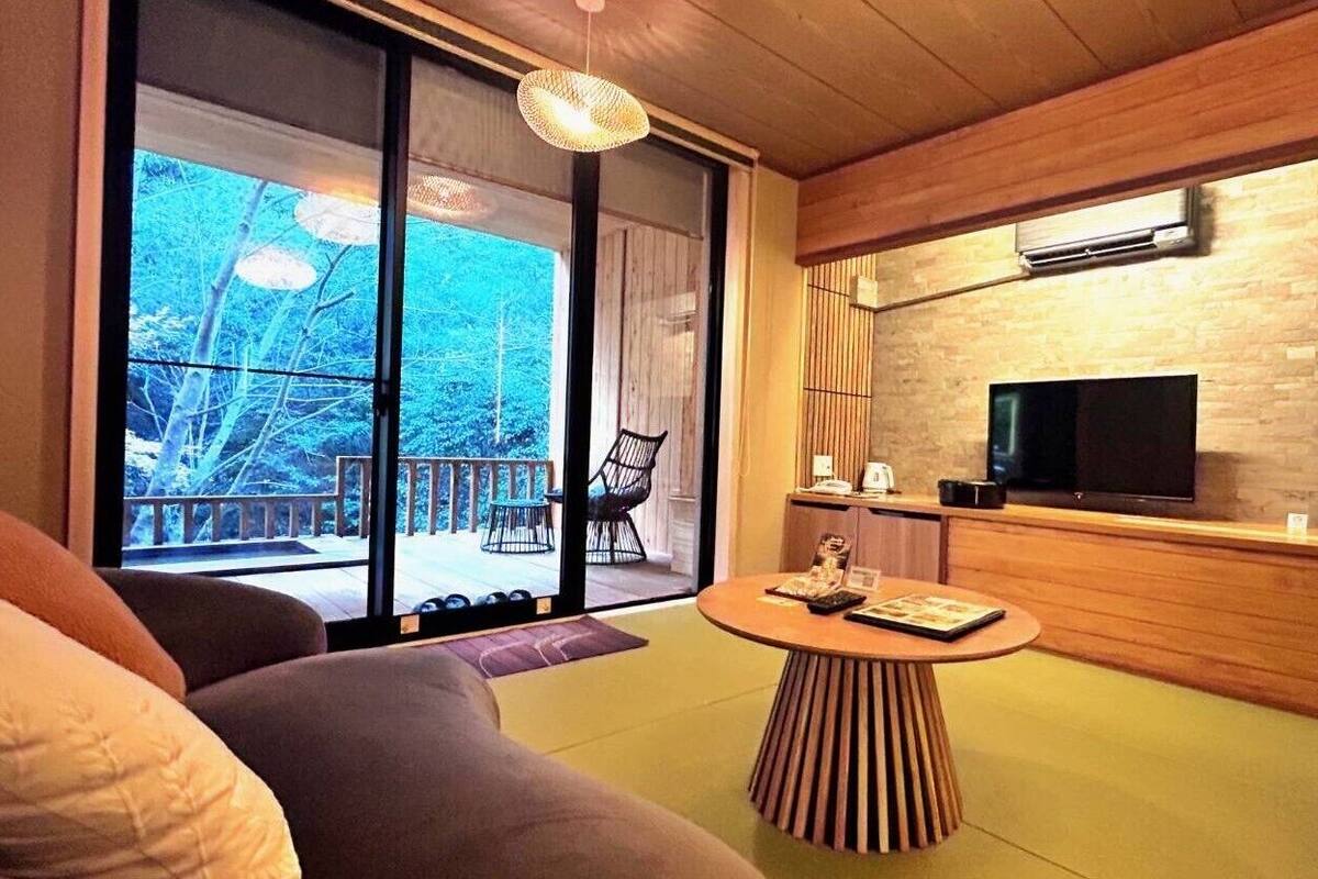Una sala de estar moderna con un amplio ventanal corredero que da a una terraza exterior, una mesa de centro redonda de madera, un televisor de pantalla plana montado en una pared de ladrillo y un sofá.