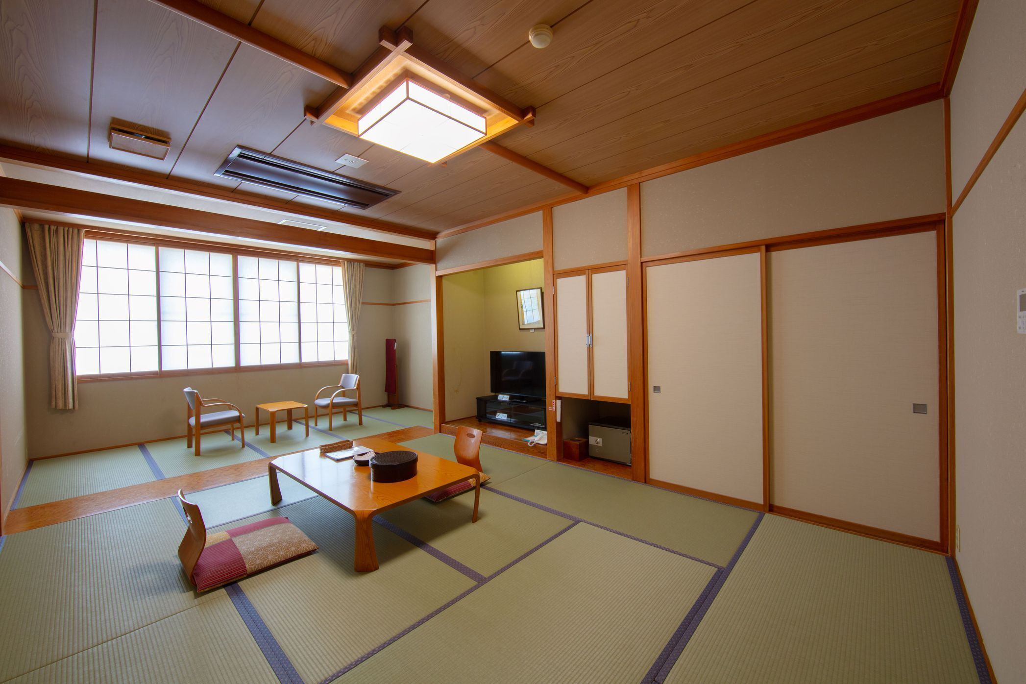 Ein traditioneller japanischer Raum mit Tatami-Bodenbelag, einem niedrigen Tisch, Stühlen, einem Fernseher und Schiebetüren.