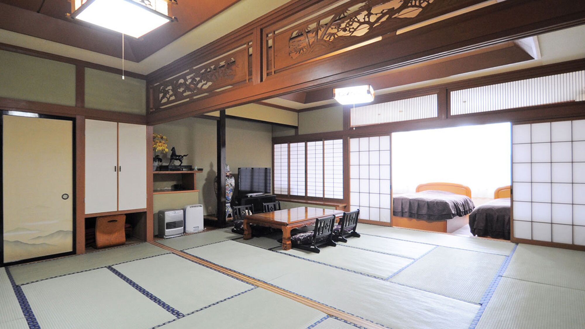 A traditional Japanese room with tatami flooring, a low table with chairs, a bed with a futon, sliding doors, and a television.