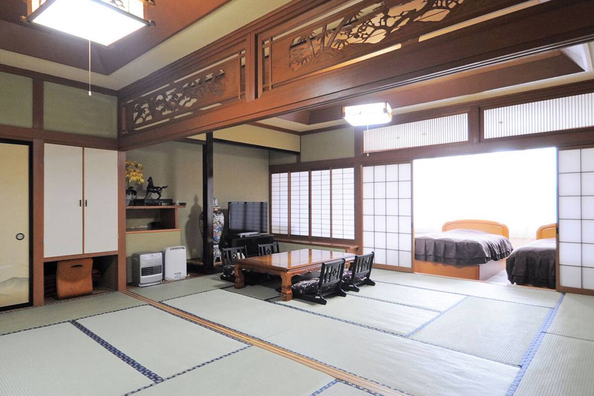A traditional Japanese room with tatami flooring, a low table with chairs, a bed with a futon, sliding doors, and a television.