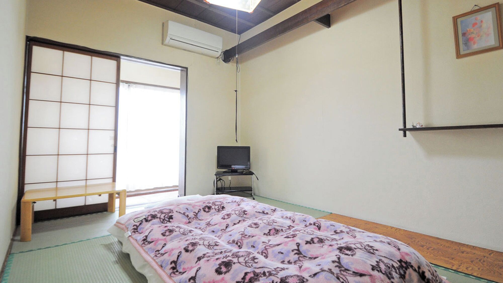 A traditional Japanese room with tatami flooring, a futon bed, a television on a stand, and a sliding door.
