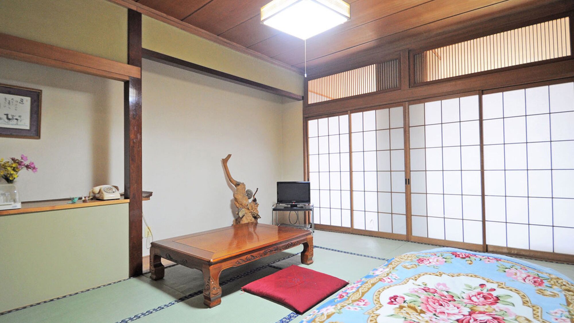 Une pièce de style japonais traditionnel, avec un sol recouvert de tatamis, une basse table en bois, un coussin rouge, un téléviseur posé sur un meuble et des paravents en papier de riz (shoji).