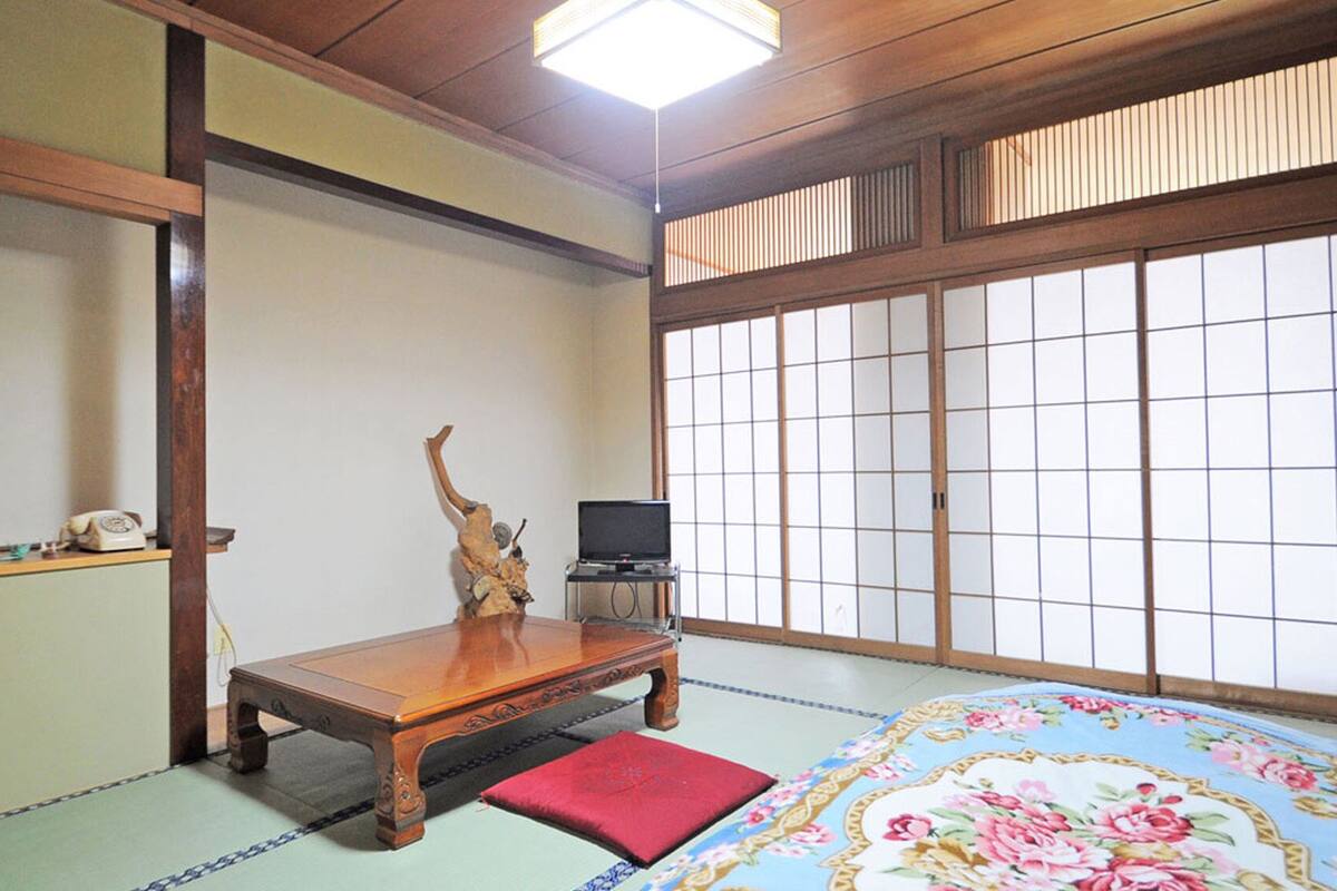 A traditional Japanese room with tatami flooring, a low wooden table, a red cushion, a television on a stand, and shoji screens.