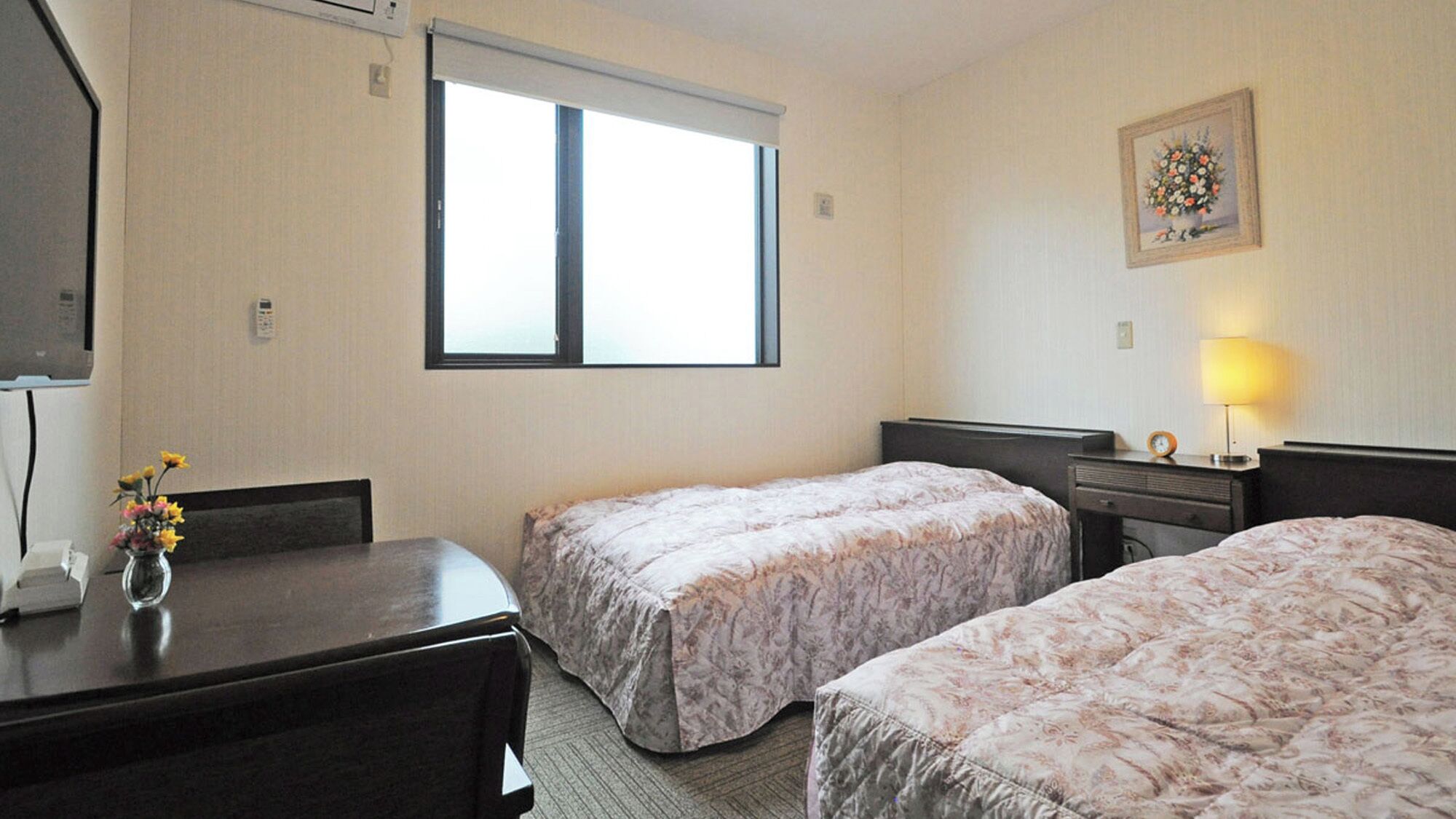 A hotel room with two beds, a desk, a chair, a window, a lamp, and a framed picture on the wall.