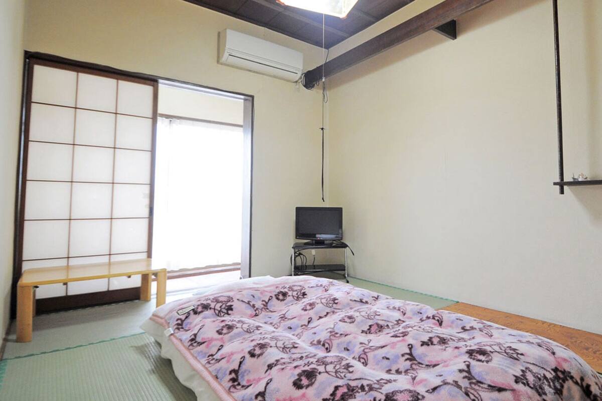 A traditional Japanese room with tatami flooring, a futon bed, a television on a stand, and a sliding shoji screen.