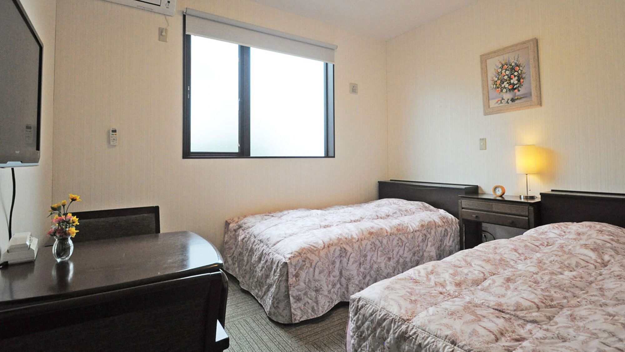 Une chambre d’hôtel avec deux lits, un bureau, une chaise, une fenêtre, une lampe et un tableau encadré au mur.