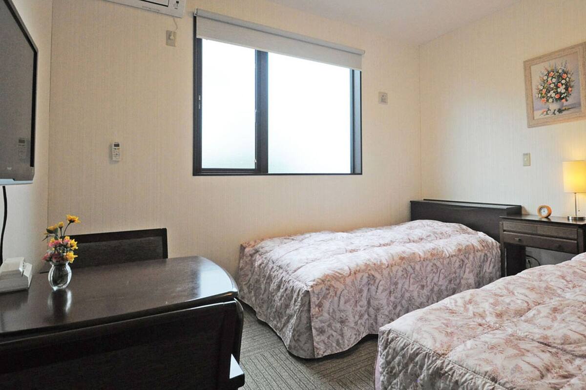 A hotel room with two beds, a desk, a chair, a window, a lamp, and a framed picture on the wall.