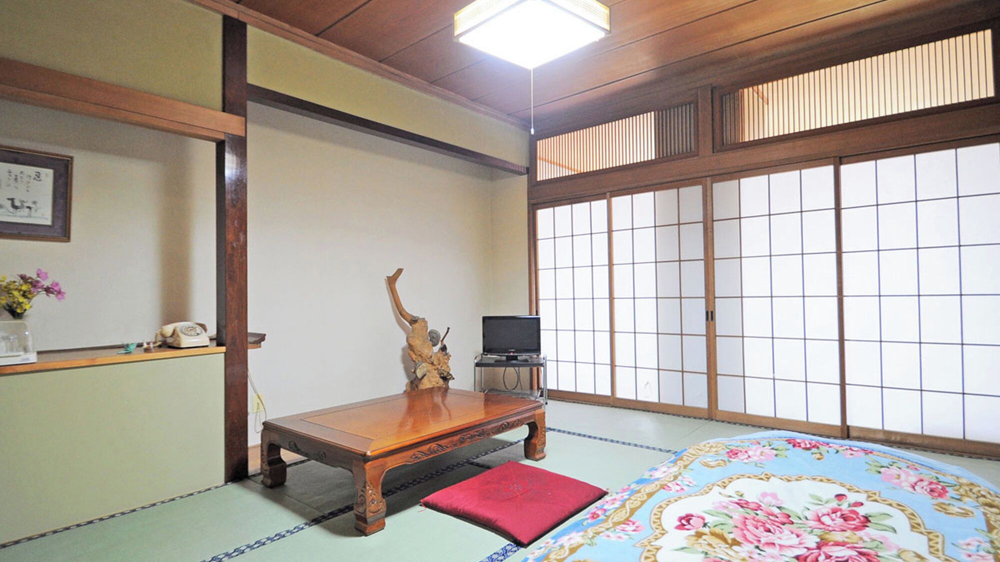 A traditional Japanese room with tatami flooring, a low wooden table, a red cushion, a television on a stand, and shoji screens.