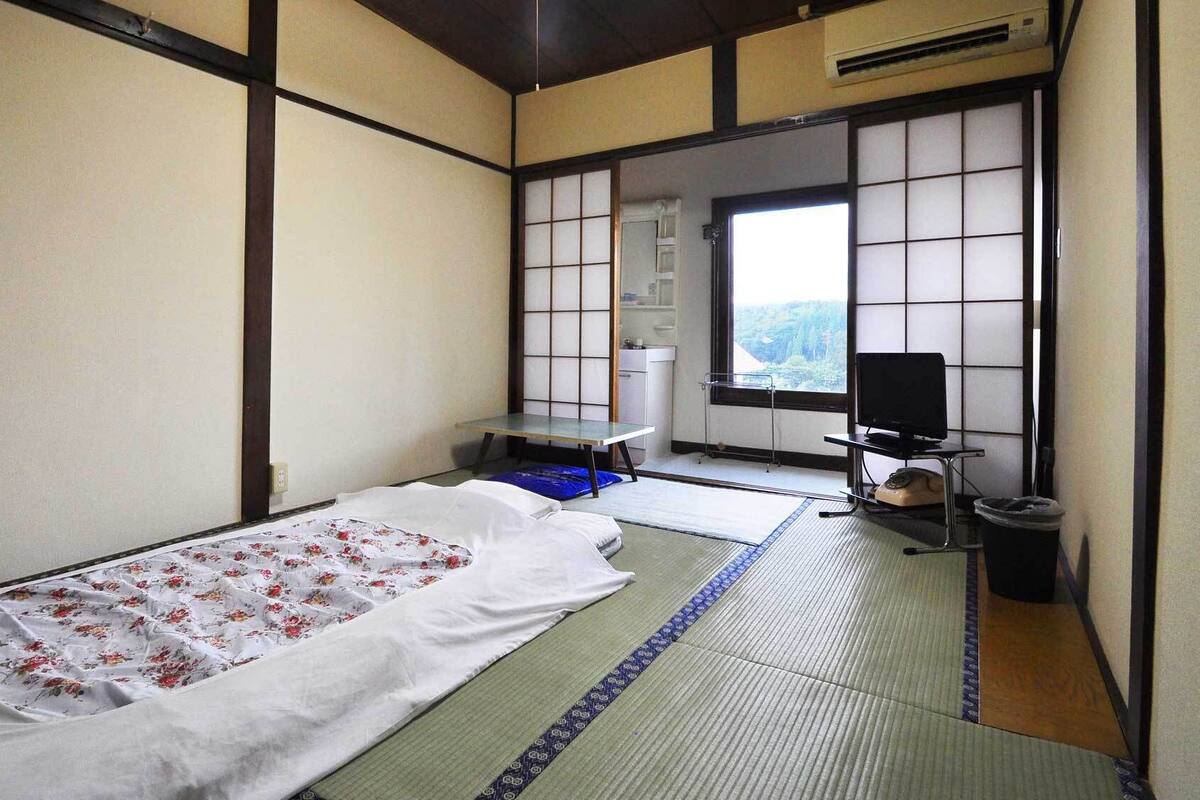 A traditional Japanese room with tatami flooring, a futon bed, a television, and a view of the outdoors.
