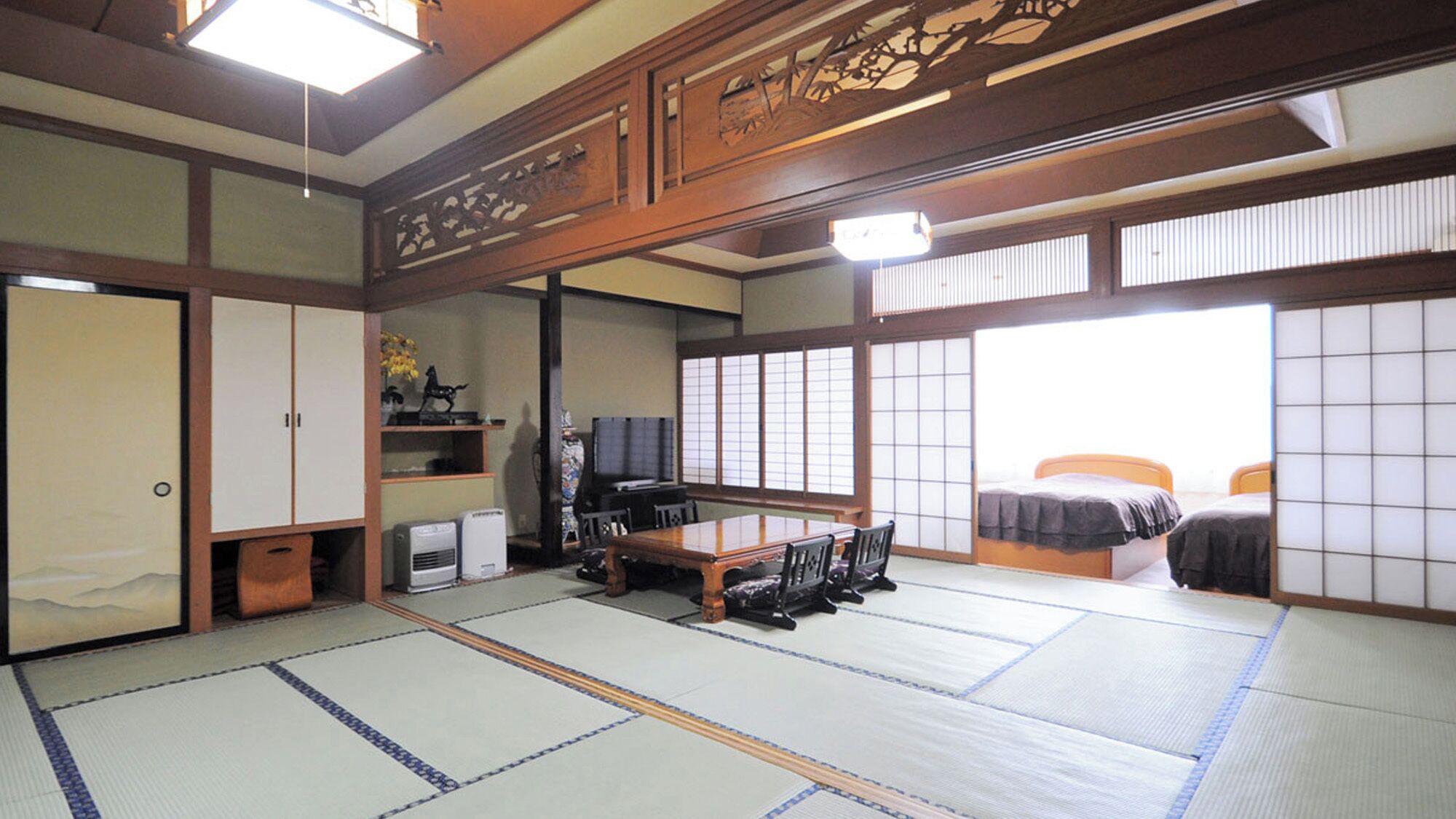 A traditional Japanese room with tatami flooring, a low table with chairs, a bed with a futon, sliding doors, and a television.