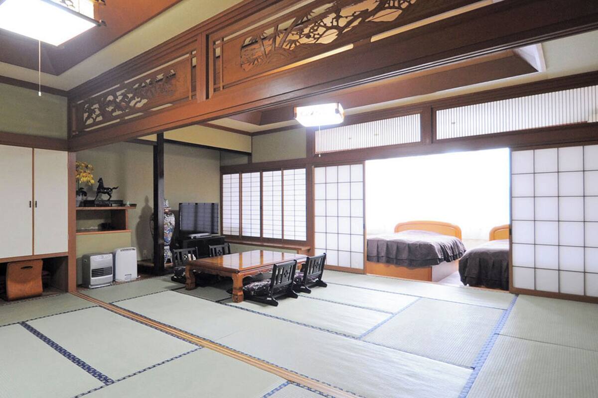 A traditional Japanese room with tatami flooring, a low table with chairs, a bed with a futon, sliding doors, and a television.