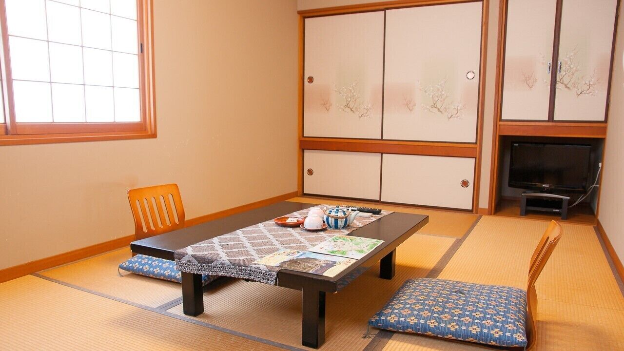 Une pièce avec un sol recouvert de tatamis, une petite table basse, des coussins, un téléviseur et des portes coulissantes.