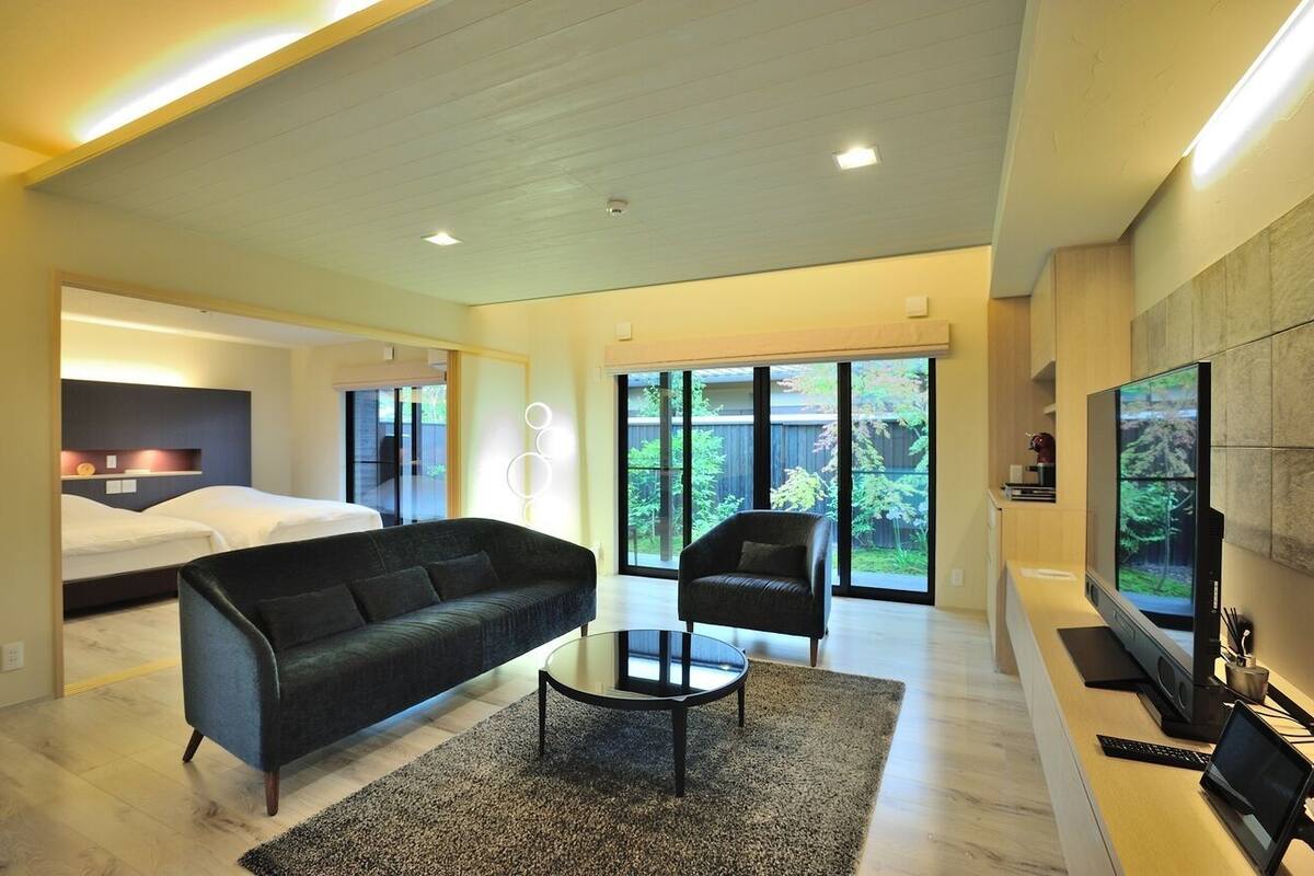 Uma sala de estar moderna com sofá, poltrona e mesa de centro, uma cama, televisão e grandes janelas com vista para o verde.