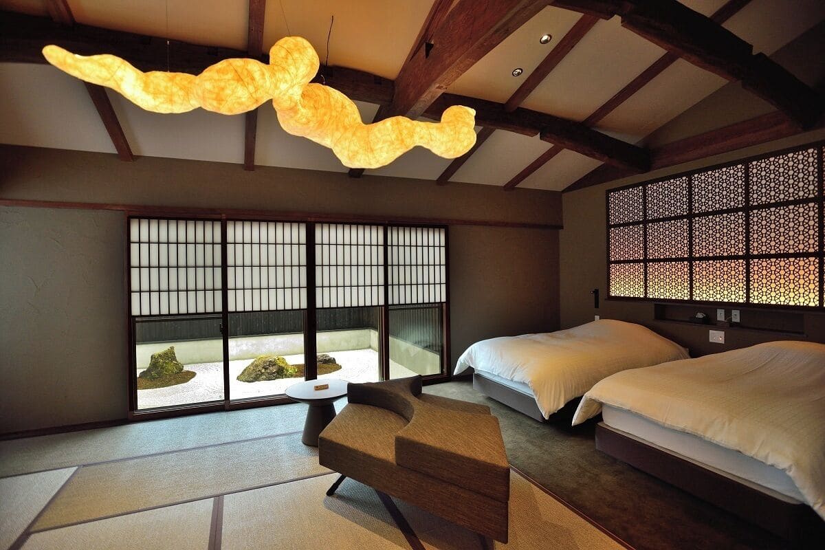 Quarto tradicional japonês com tatames, cama de madeira baixa, um banco acolchoado e uma grande janela com vista para pedras.