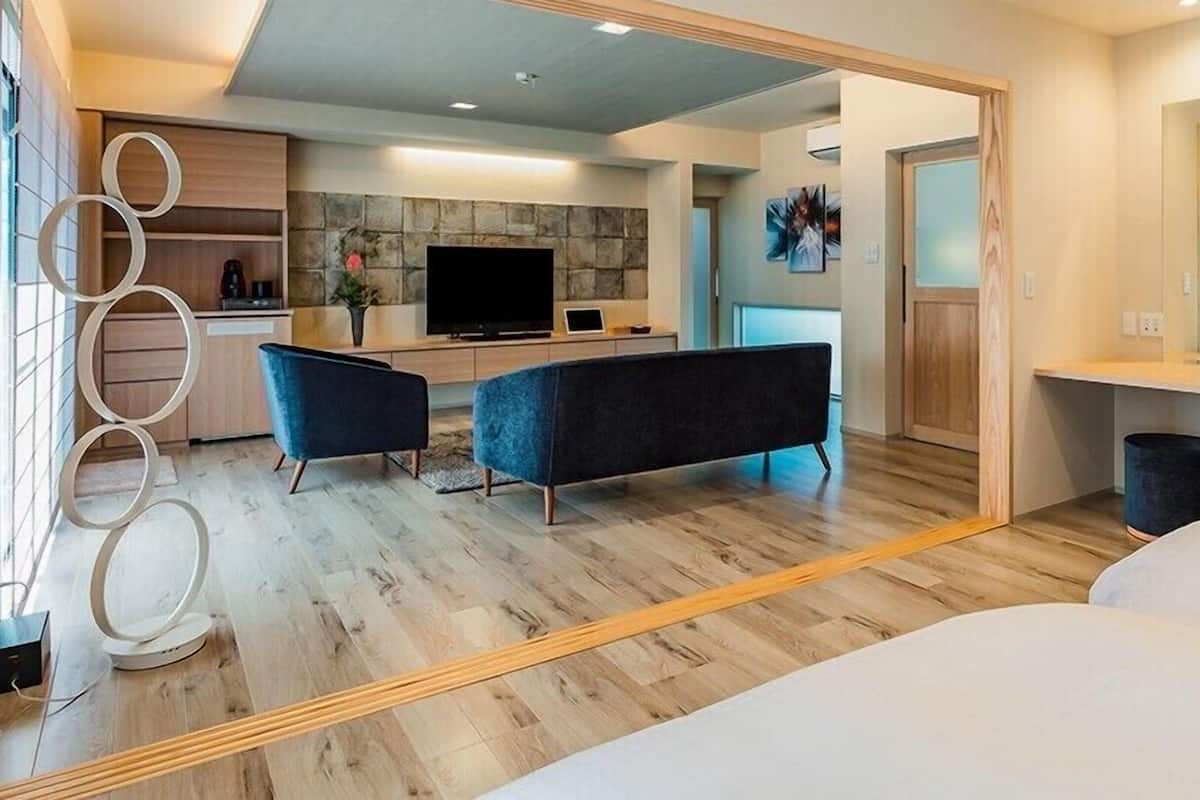 Quarto de hotel moderno com uma cama grande, um espaço de estar com sofá e poltronas, uma área de jantar com TV e uma parede de destaque em pedra, e um banheiro com espelho e pia.