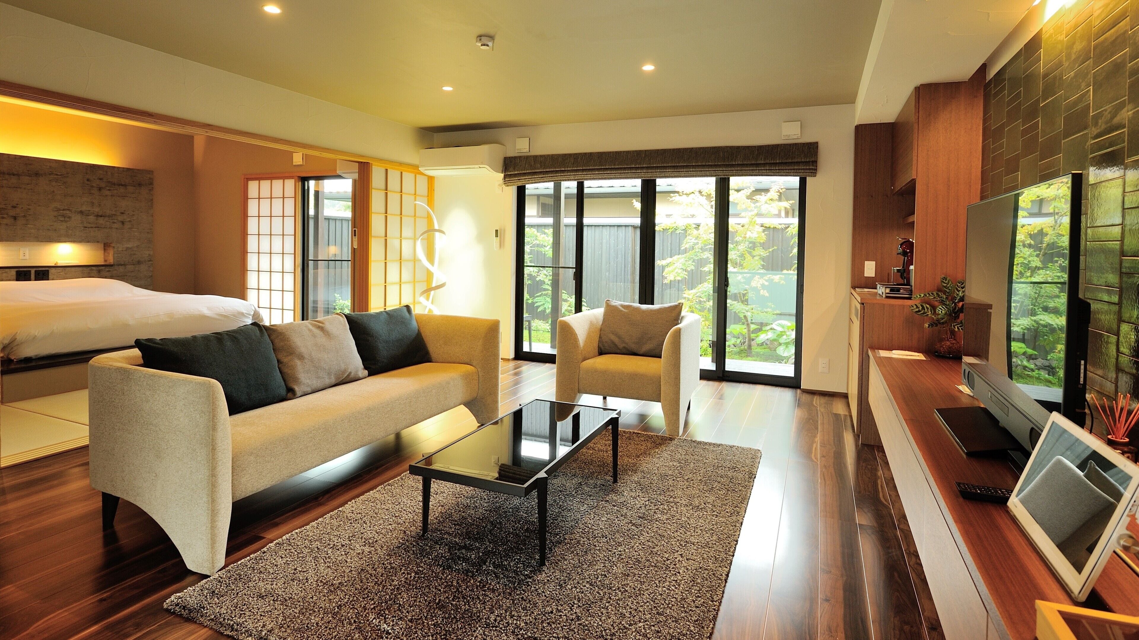 A modern hotel room with a sofa, armchair, flat-screen TV, and a large window offering a view of greenery.