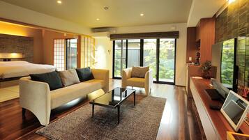 A modern hotel room with a sofa, armchair, flat-screen TV, and a large window offering a view of greenery.