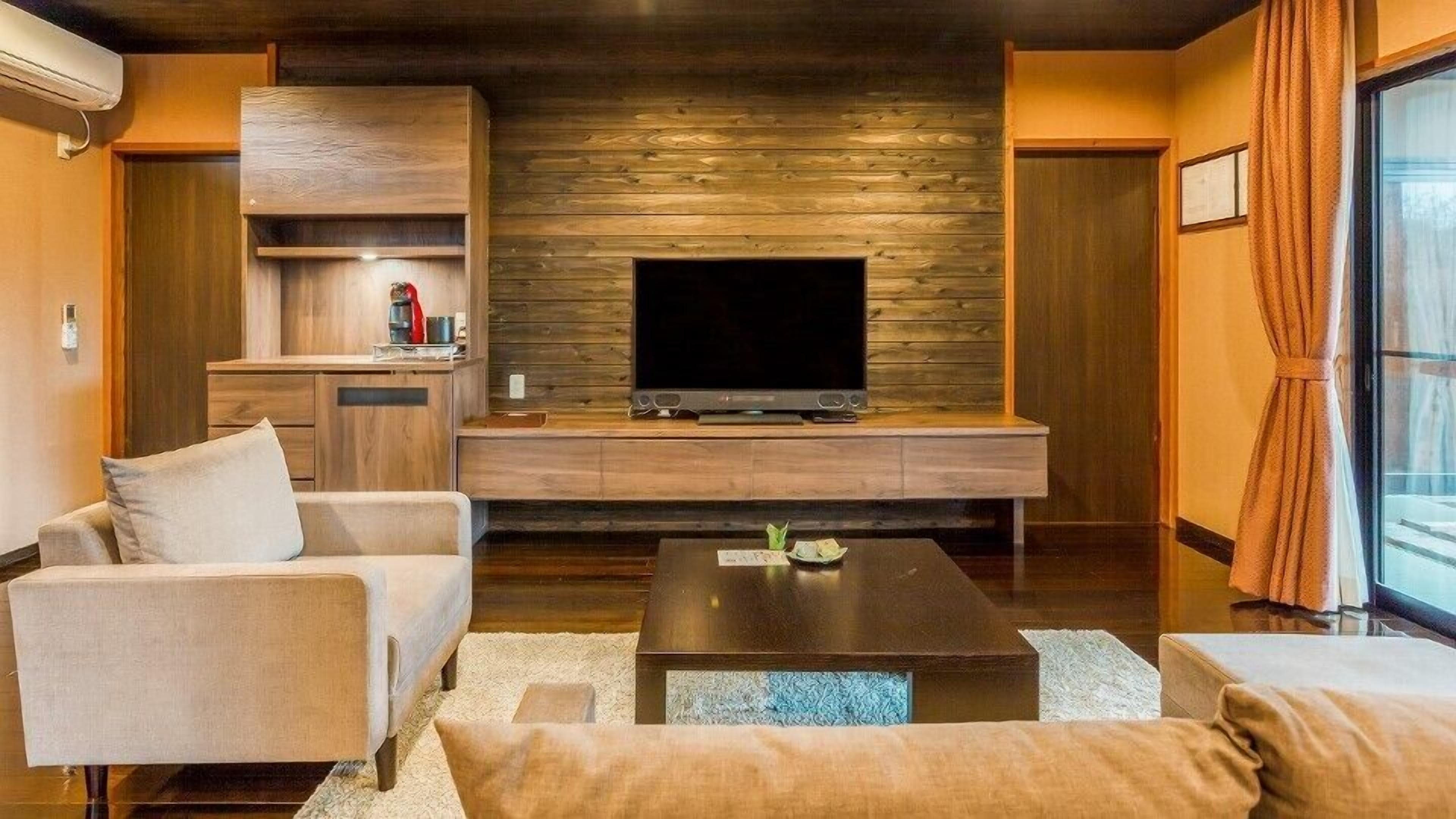 A modern living room with a flat-screen TV, wooden wall paneling, a kitchen area, and a large window with curtains.