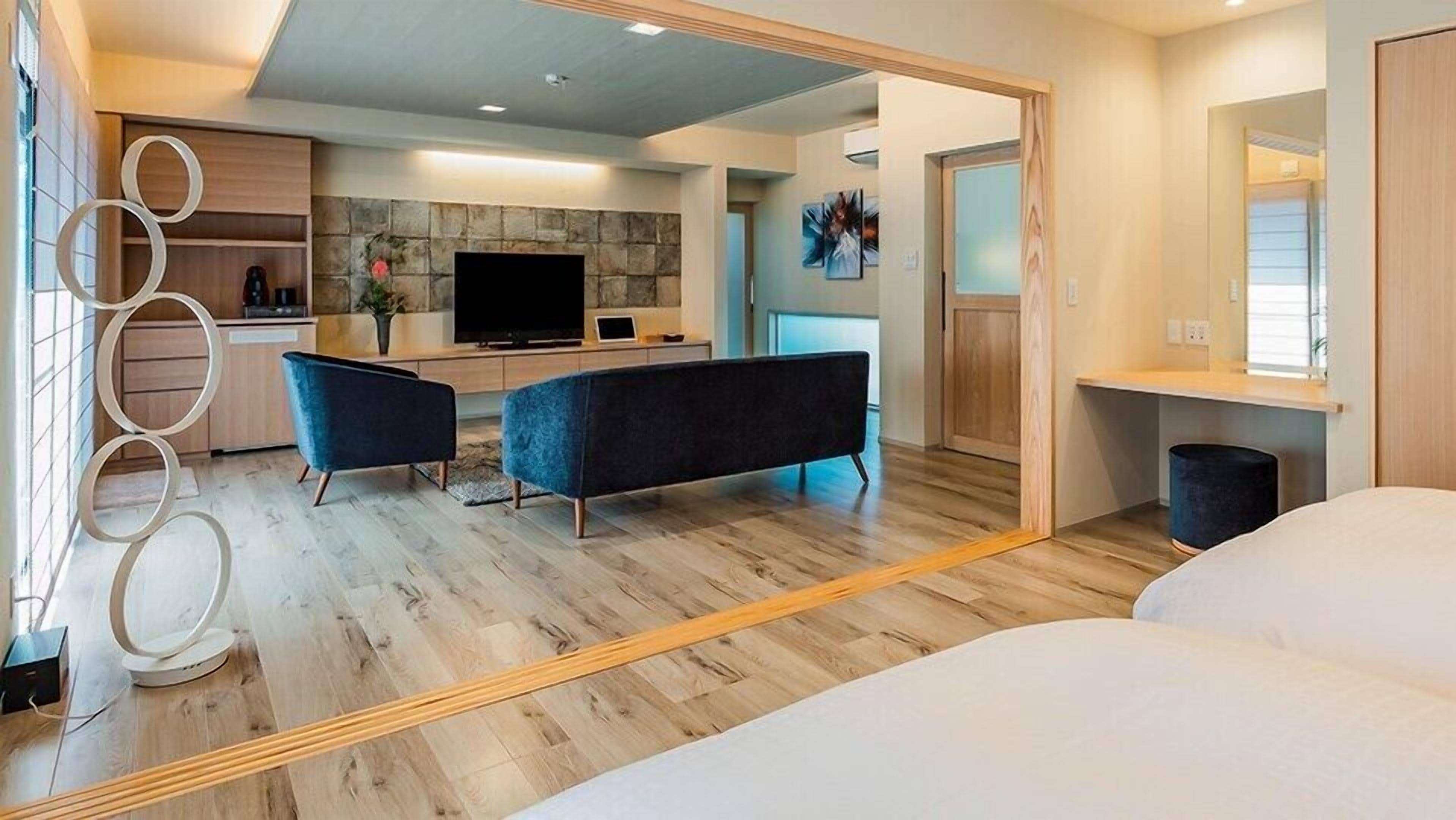 A modern hotel room with a large bed, a sitting area with a sofa and armchairs, a dining area with a TV and a stone accent wall, and a bathroom with a mirror and sink.