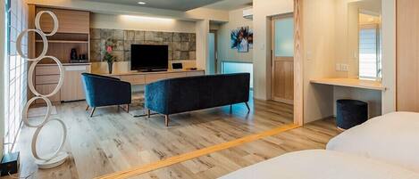 Una habitación de hotel moderna con una cama grande, una zona de estar con sofá y sillones, un comedor con televisor y una pared de acento de piedra, y un baño con espejo y lavamanos.