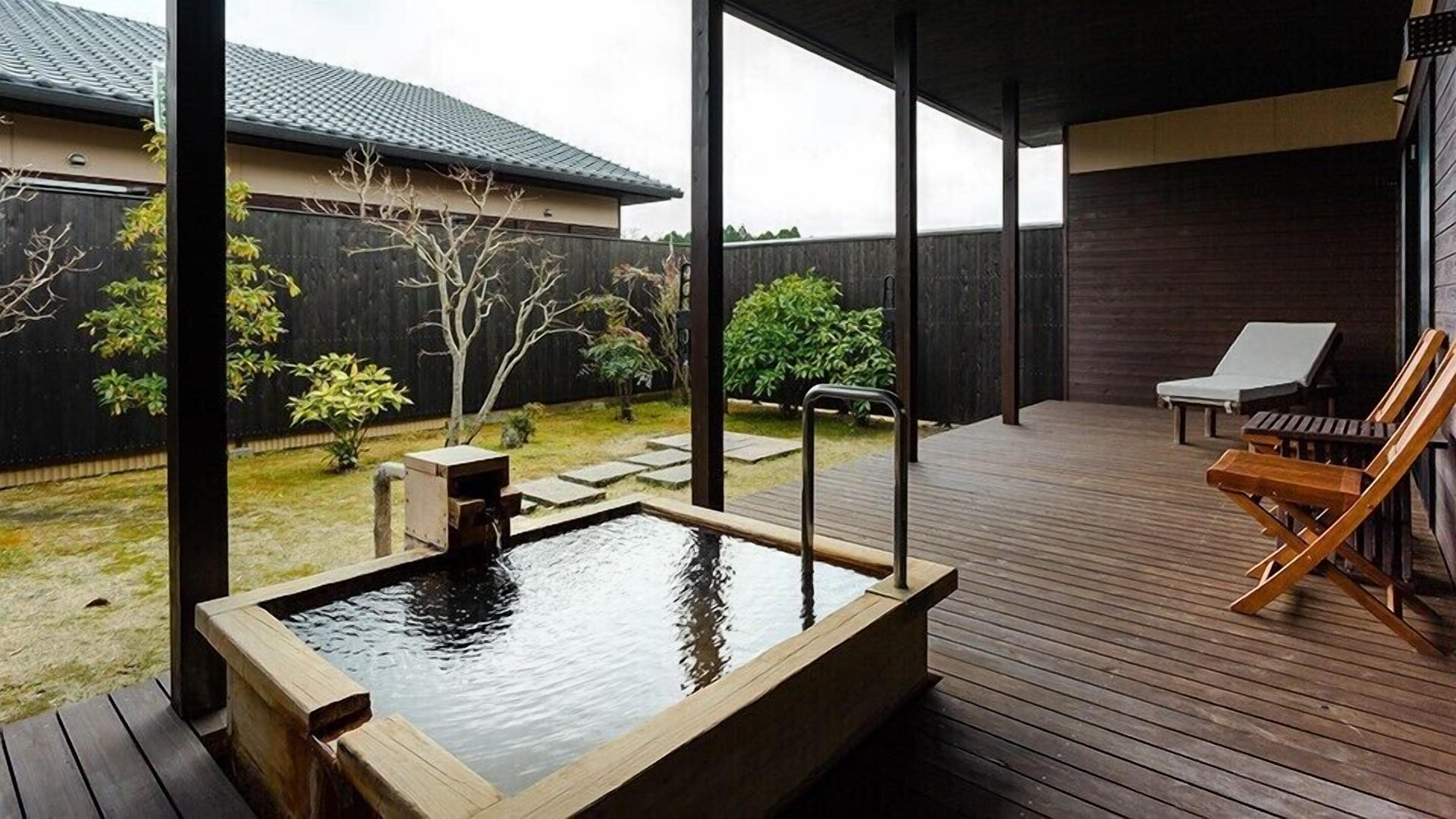 A wooden deck with a hot tub, lounge chairs, and a view of a house with a dark roof.