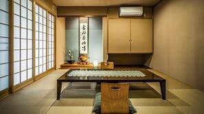 Una tradizionale stanza giapponese con pavimento in tatami, un tavolino con un sottotavolo di pizzo, una sedia di legno, un paravento shoji, un condizionatore d'aria a parete e un rotolo di calligrafia.