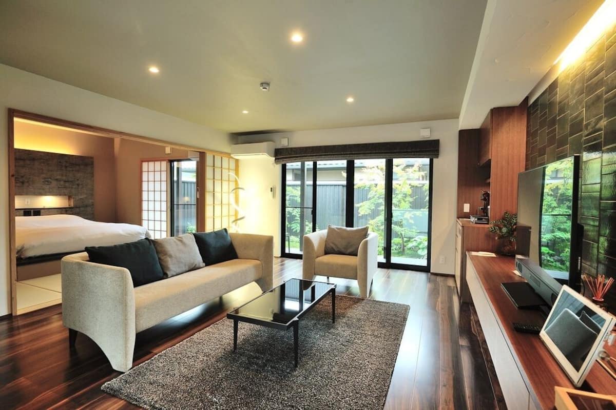 Uma sala de estar moderna com sofá, poltrona e mesa de centro, uma TV e uma janela grande com vista para o verde.