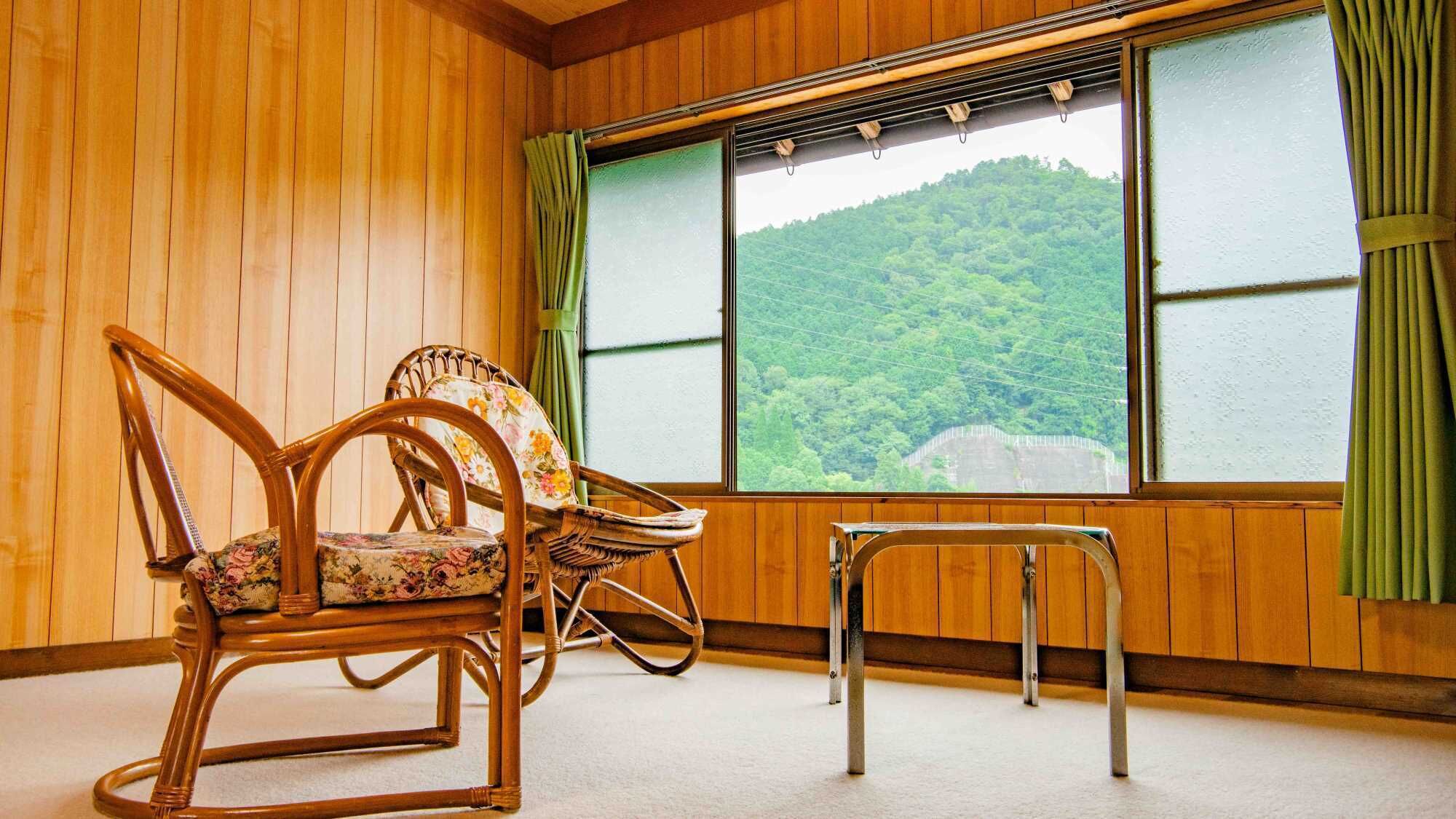 Una habitación con paredes de madera, dos sillones de mimbre con tapicería floral, una mesita y un ventanal con vistas a una colina boscosa.
