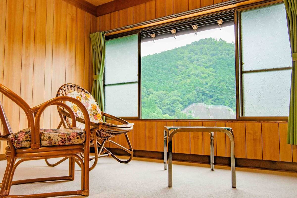 A room with wooden walls, two wicker chairs with floral upholstery, a small table, and a large window with a view of a forested hill.